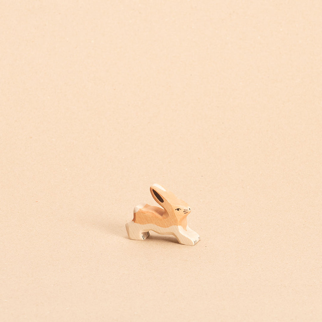 Ein hellbrauner, stehender Hase aus Holz mit weißem Bauchfell, weißer Blume, schaut nach vorne links. Seine Ohren sind nach oben hin gespitzt.