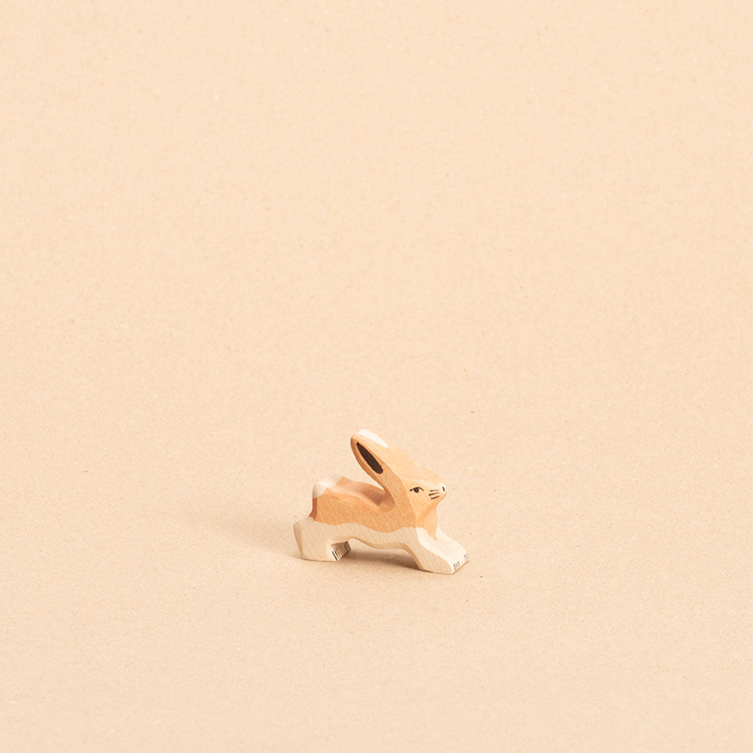 Ein hellbrauner, stehender Hase aus Holz mit weißem Bauchfell, weißer Blume, schaut nach vorne links. Seine Ohren sind nach oben hin gespitzt.