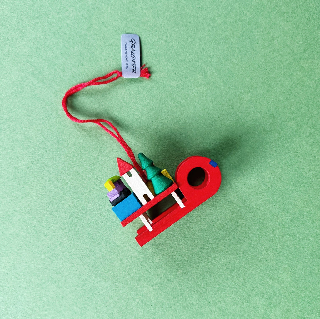 Graupner Baumschmuck Schlitten mit Geschenken ist ein kleiner roter Schlitten aus Holz, mit niedlichen Minigeschenken beladen. Auch ein winziges Tannenbäumchen und ein kleines Häuschen sind dabei. Mit dem roten Bändchen kann der Anhänger an einem Zweig befestigt werden.