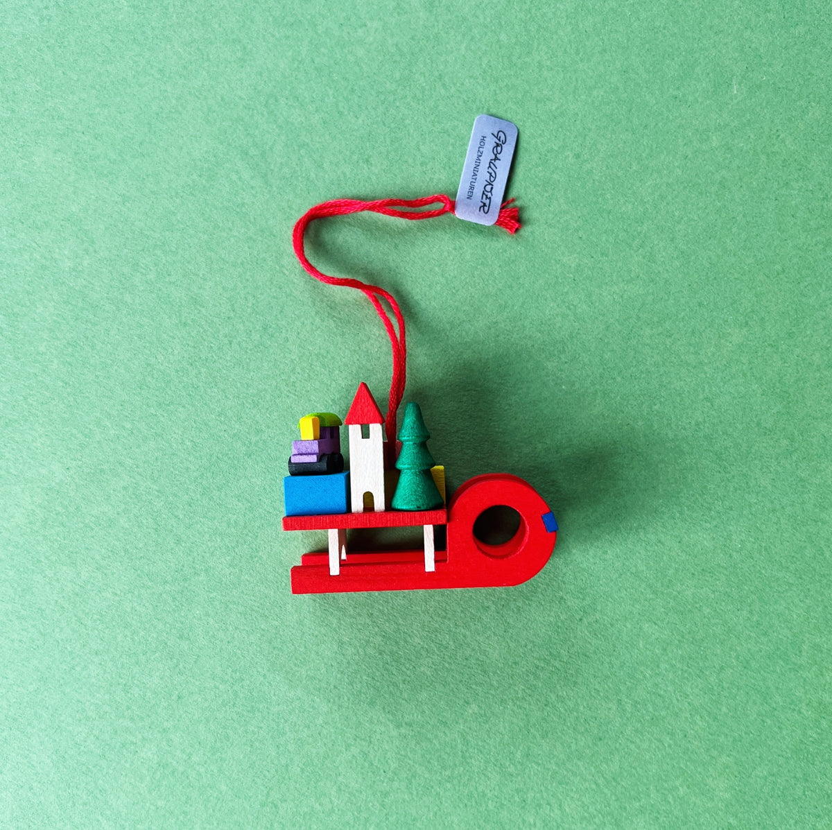 Graupner Baumschmuck Schlitten mit Geschenken ist ein kleiner roter Schlitten aus Holz, mit niedlichen Minigeschenken beladen. Auch ein winziges Tannenbäumchen und ein kleines Häuschen sind dabei. Mit dem roten Bändchen kann der Anhänger an einem Zweig befestigt werden.