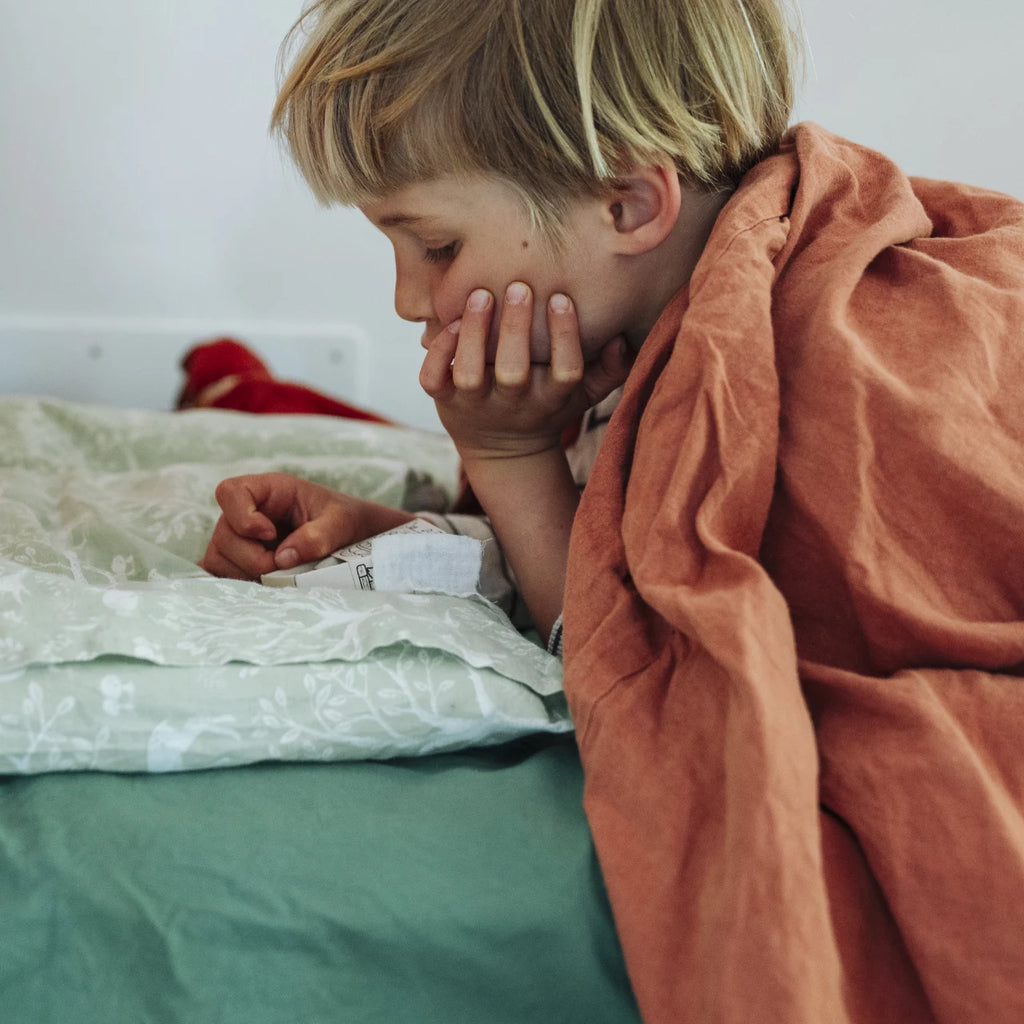 Die Adventszeit wird durch den Grapat Adventskalender 2026 so richtig gemütlich. Ein Kind liegt auf einem grün bezogenen Bett und ist in eine rote Decke eingekuschelt. Das Kind ist im Profil zu sehen und schaut verträumt auf eine der kleinen Schachteln. Es bewundert die darin enthaltenen kleinen Schätze, die für den Betrachter verborgen bleiben – genauso geheimnisvoll, wie es für einen Adventskalender typisch ist.