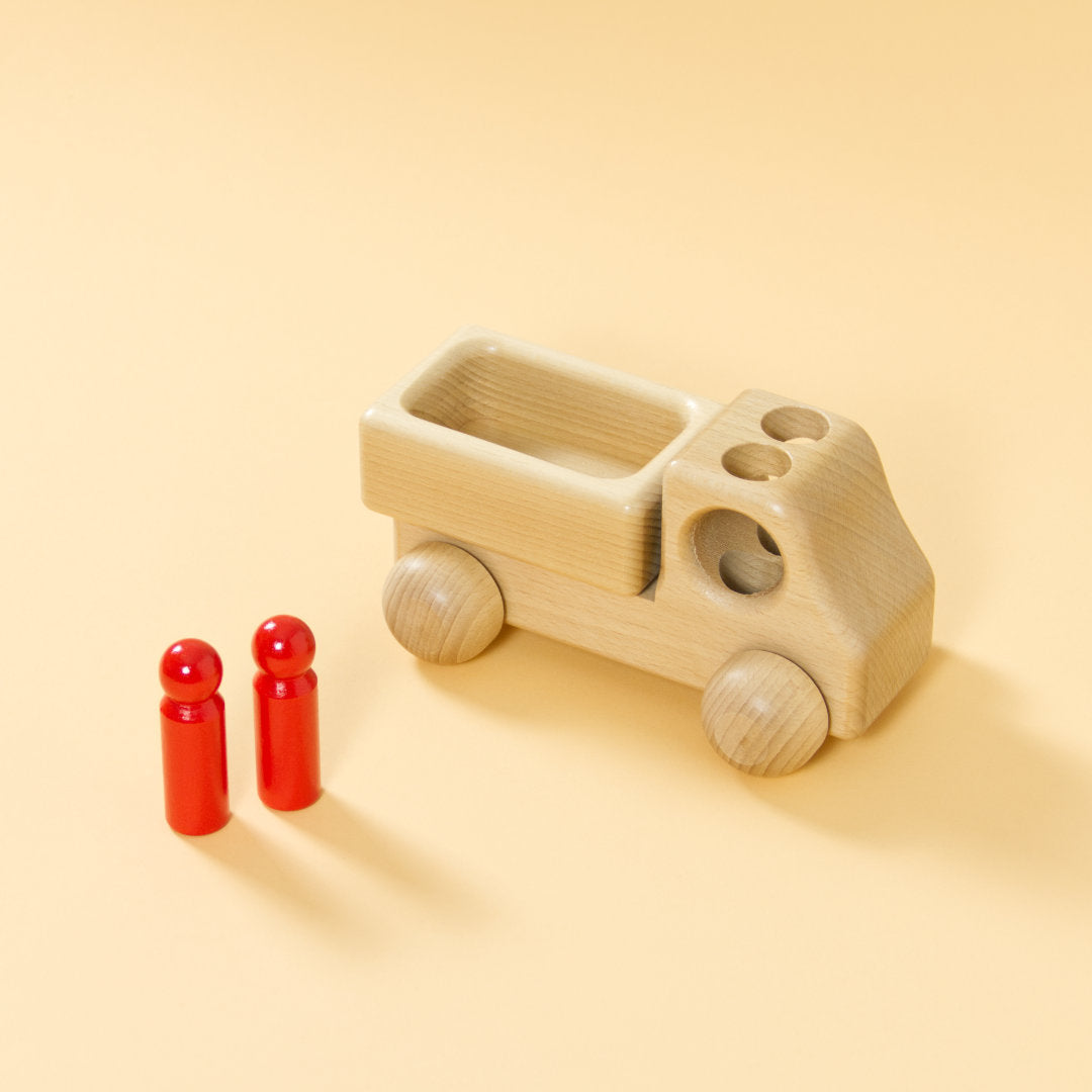 Lieferwagen aus Buchenholz, natur, mit abgerundeter Formensprache. Offene Ladefläche, Fahrerkabine mit zwei kegelförmigen, roten Figuren, die Figuren stehen, über Löcher im Dach der Fahrerkabine können sie einsteigen.