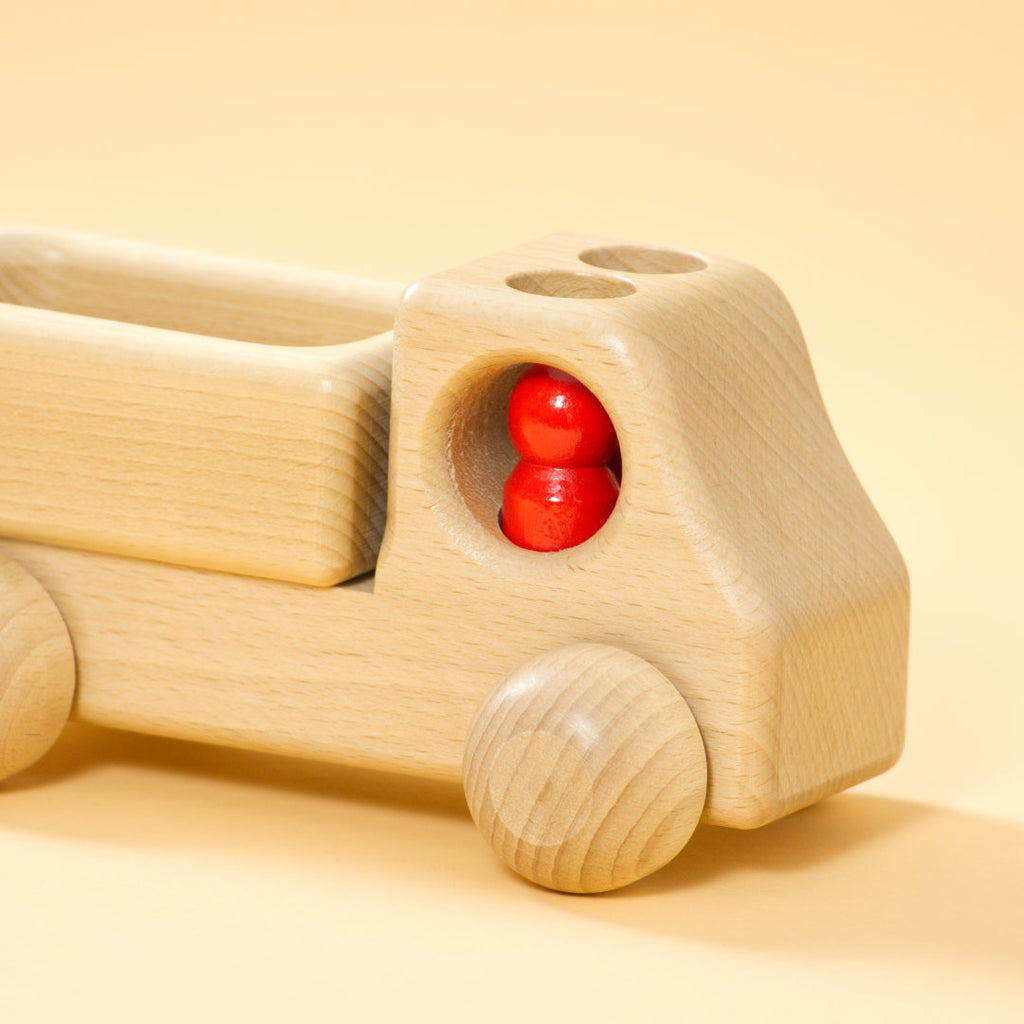 Lieferwagen aus Buchenholz, natur, mit abgerundeter Formensprache. Offene Ladefläche, Fahrerkabine mit zwei kegelförmigen, roten Figuren.