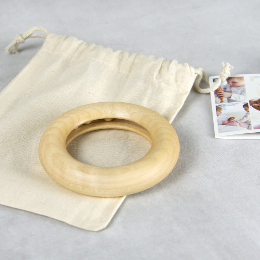 Fotografiertes Arrangement mit dem Greifling mit Innenbahn und 3 darin liegenden Kugeln. Der Greifling ist aus Naturholz von Kügelhaus gefertigt und ist auf einem einfarbig, beige Jutebeutel fotografiert mit Zugband. Rechts daneben im Anschnitt ist ein collagierter Fotoausdruck als Karte zu sehen.
