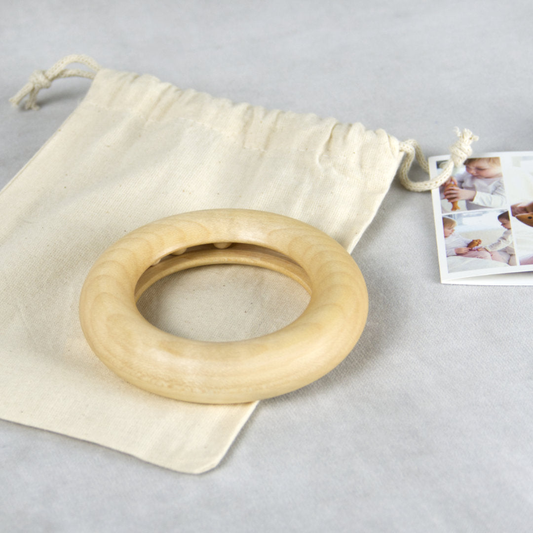 Fotografiertes Arrangement mit dem Greifling mit Innenbahn und 3 darin liegenden Kugeln. Der Greifling ist aus Naturholz von Kügelhaus gefertigt und ist auf einem einfarbig, beige Jutebeutel fotografiert mit Zugband. Rechts daneben im Anschnitt ist ein collagierter Fotoausdruck als Karte zu sehen.