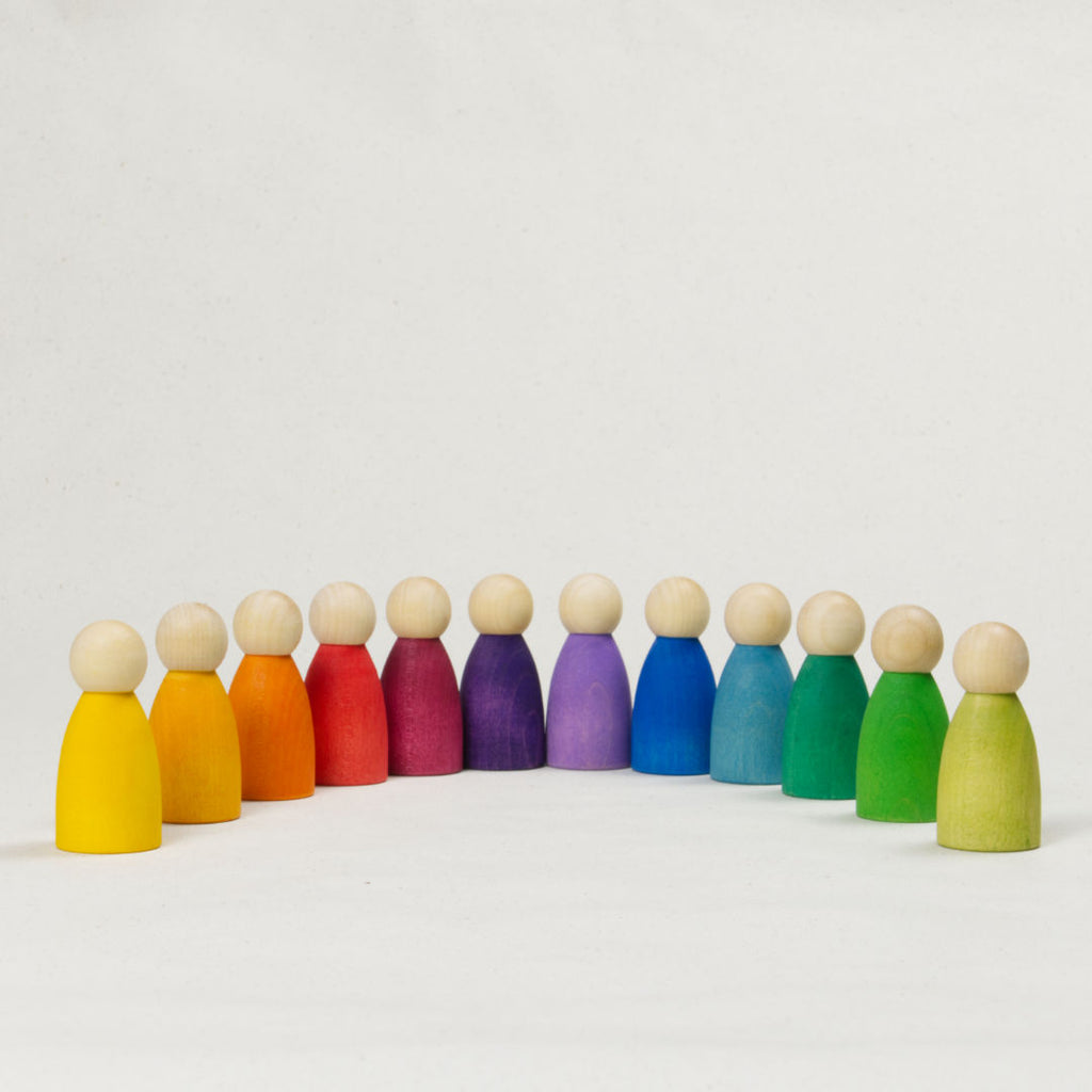 Zwölf gleichgeformte Holzfiguren mit Köpfen in Naturholz und unterschiedlich farbigen Körpern, als Regenbogen angeordnet.