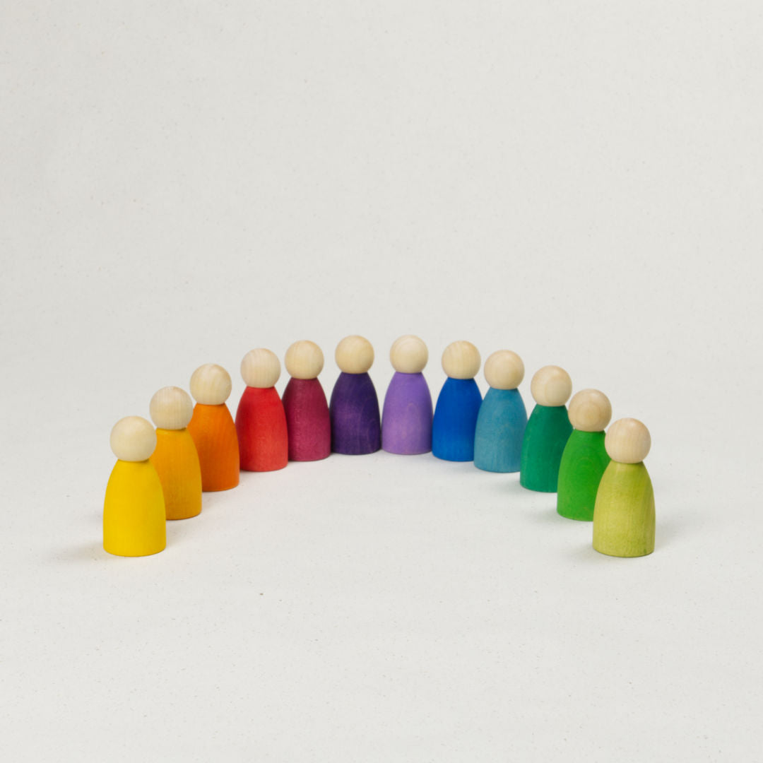 Zwölf gleichgeformte Holzfiguren mit Köpfen in Naturholz und unterschiedlich farbigen Körpern, als Regenbogen angeordnet.
