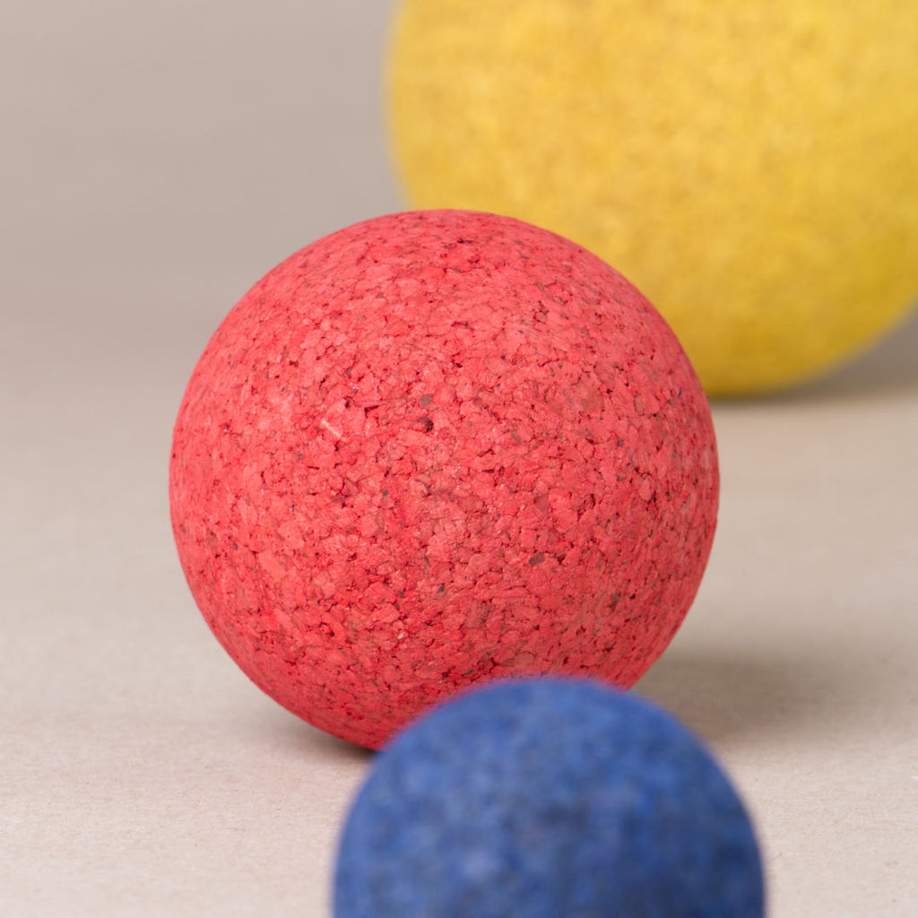 Komposition aus drei Kork Kugeln in abgestufter Größe von Korxx, die kleinste in blau, die Mittlere in rot und die Größte in gelb. Im Mittelpunkt scharf zu sehen ist die rote Kugel.