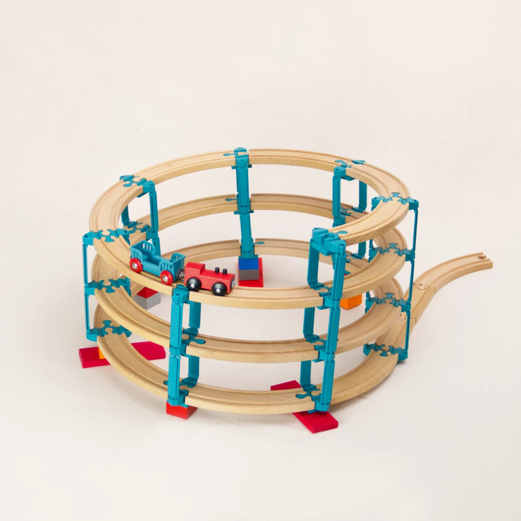 Das Adapter-Set Helix 3 von Yamba ermöglicht, Klemmbausteine (DUPLO®, BioBUDDi®, Hubelino® usw.) und Schienen (BRIO®, IKEA®, Eichhorn®, Hape® usw.) zu einem Säulensystem mit bis zu vier Etagen zu verbinden. Auf der Bahn fährt eine rote Holzlokomotive nach unten, an der ein türkisfarbener Wagen hängt.