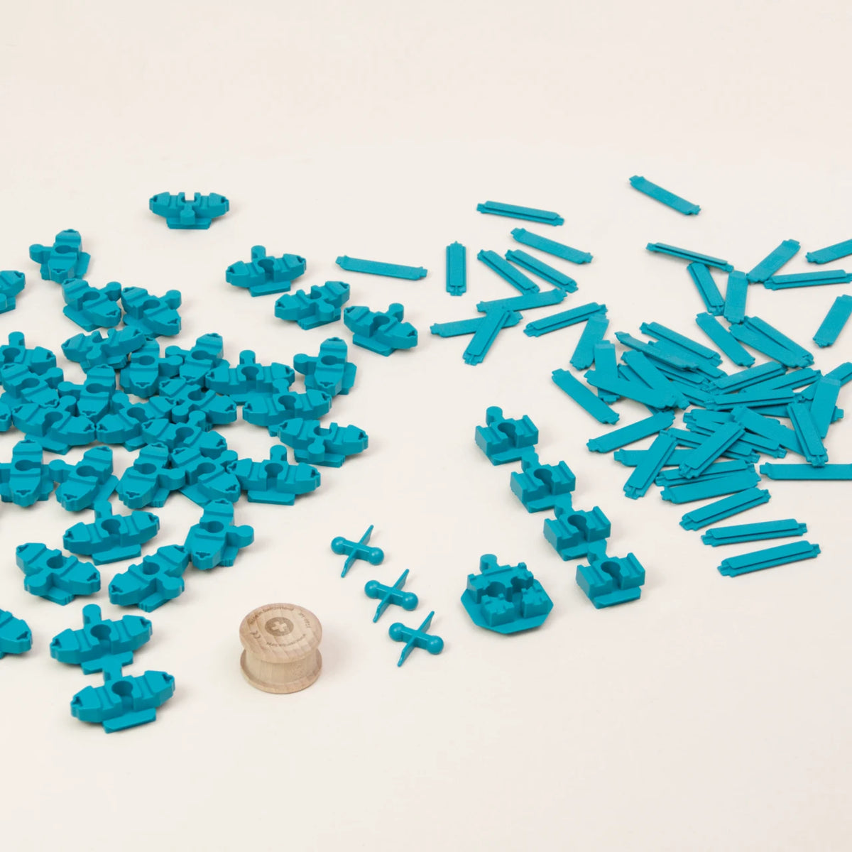 Zu sehen sind alle Elemente des Adapter-Sets Helix 5 von Yamba: insgesamt 104 türkise Bausteine in fünf unterschiedlichen Formen: Verbindungsstück Link, Verbindungsstück LinkUp, Verbindungsknochen Bar, Verbindungsstück Cross und Säule Lift. Die Einzelteile bestehen aus hochwertigem ABS-Kunststoff und sind im Spritzgussverfahren hergestellt. Zudem enthält das Set einen Rolli, eine zylindrische Holzwalze.