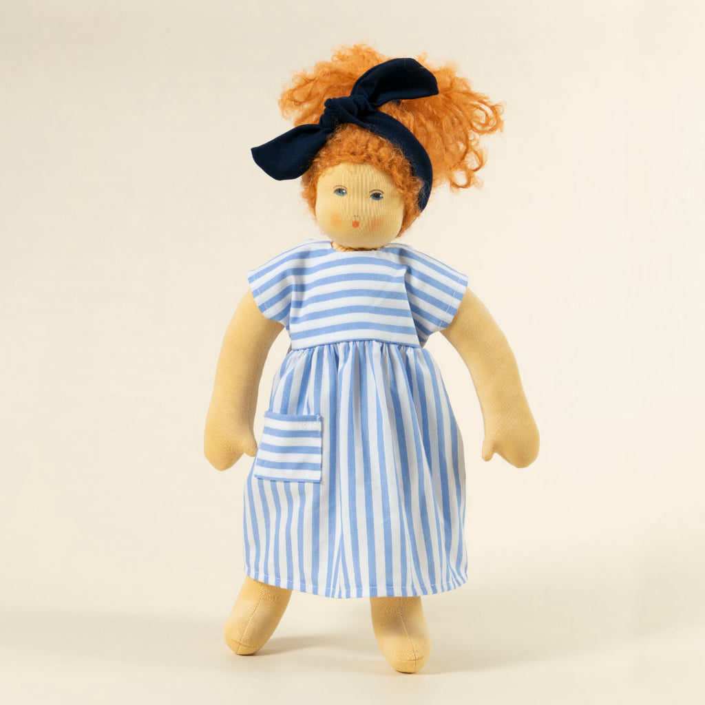 Nanchen Puppe mit heller Haut und roten Haaren stehend. Sie trägt eine blaue Haarschleife und ein blau-weiß gestreiftes Kleid.