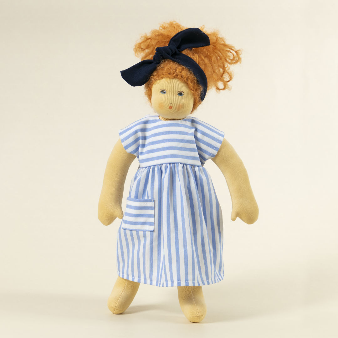 Nanchen Puppe mit heller Haut und roten Haaren stehend. Sie trägt eine blaue Haarschleife und ein blau-weiß gestreiftes Kleid.