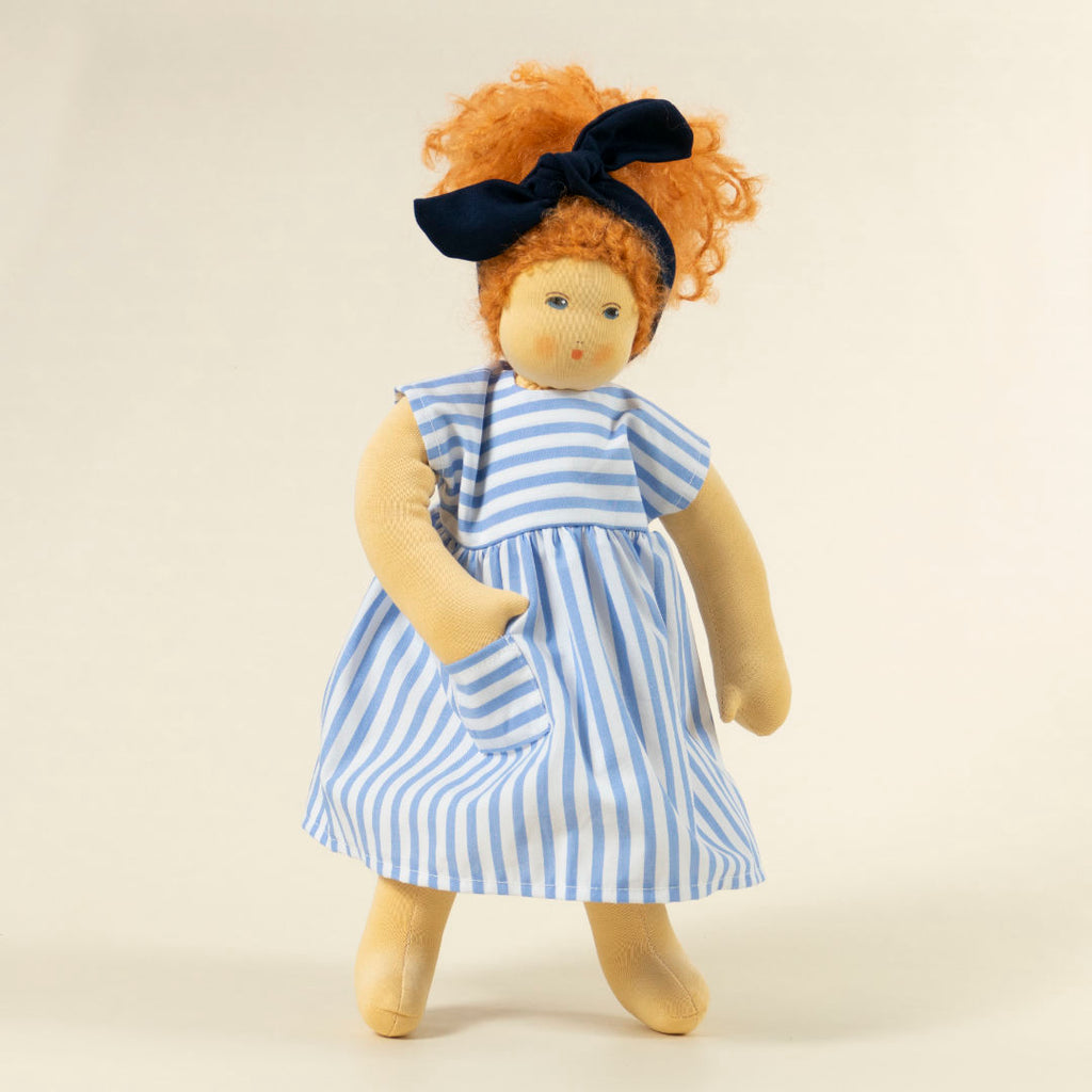 Nanchen Puppe mit heller Haut und roten Haaren stehend. Sie trägt eine blaue Haarschleife und ein blau-weiß gestreiftes Kleid.