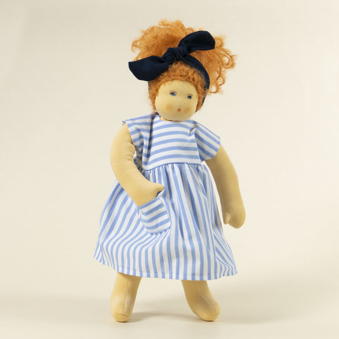 Nanchen Puppe mit heller Haut und roten Haaren stehend. Sie trägt eine blaue Haarschleife und ein blau-weiß gestreiftes Kleid.