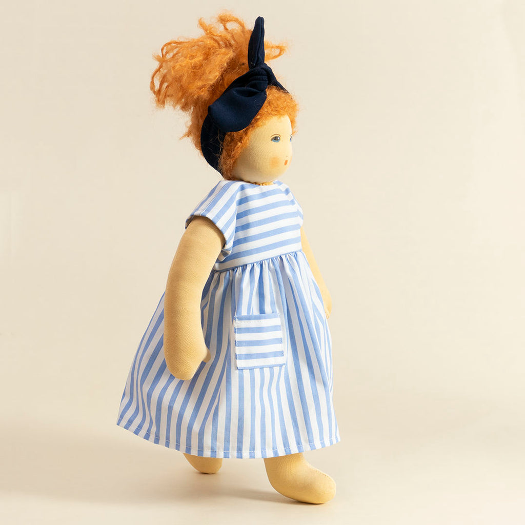 Nanchen Puppe mit heller Haut und roten Haaren stehend. Sie trägt eine blaue Haarschleife und ein blau-weiß gestreiftes Kleid.
