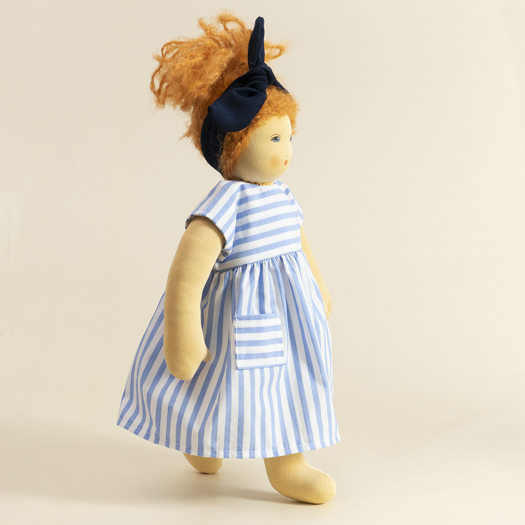 Nanchen Puppe mit heller Haut und roten Haaren stehend. Sie trägt eine blaue Haarschleife und ein blau-weiß gestreiftes Kleid.