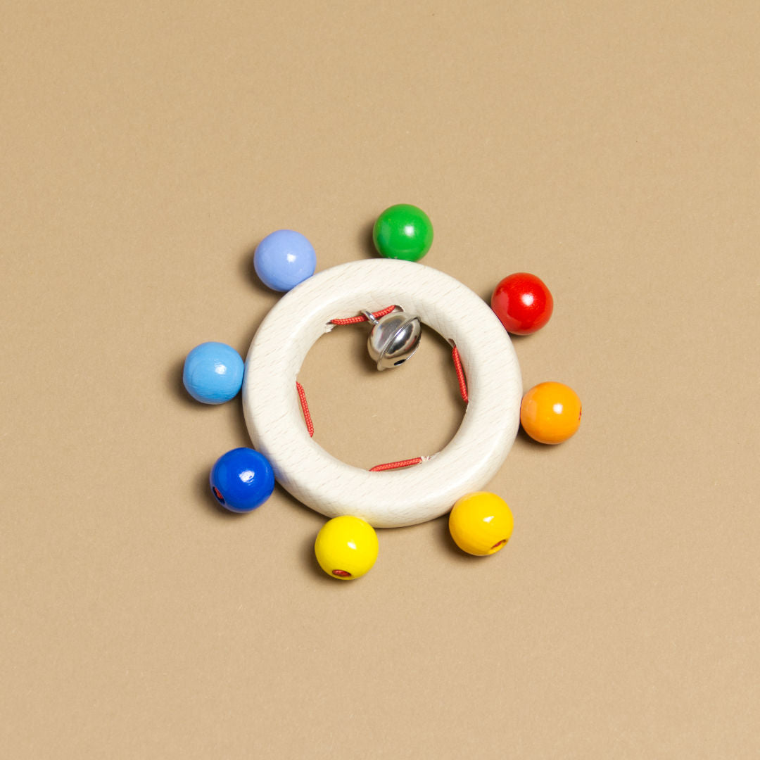Greifling mit Regenbogenperlen, bestehend aus einem naturfarbenen Ring, der am äußeren Ringrand sich befindende bunten Perlen hat, diese immer mit gleichem Abstand angeordnet, aufweist. Im Ring Innenrand befindet sich eine in 4 Segmente eingeteilte rote Schnur. An einer dieser Segmente hängt eine metallene Glocke.