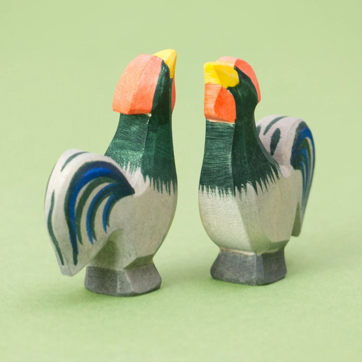 Zwei hölzerne Hähne der Marke Ostheimer. Beide Vögel stehen parallel zueinander; einer blickt nach vorne, der andere nach hinten. Das Bild verdeutlicht, dass es sich um flach ausgesägte Figuren handelt. Die Figur zeigt die Silhouette eines krähenden Hahns, die Kanten sind leicht abgerundet. Der Hahn hat einen grau grünes Gefieder mit blauen Schwanzfedern. Er hat einen gelben Schnabel und einen roten Kamm, die Augen bestehen aus kleinen schwarzen Punkten.