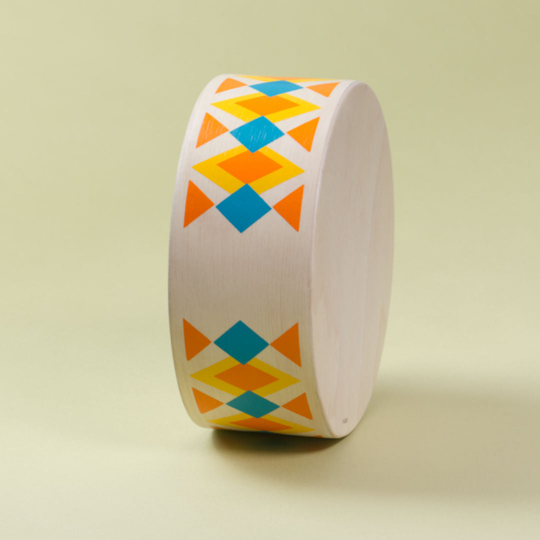 Seitenansicht ohne Loch der runden Handtrommel von Plan Toys. Das helle Holz ziert ein orange, blau, gelbes Rauten bzw. Dreiecksmuster.