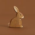 Ein brauner, sitzender Hase im Profil nach rechts schauend, ist von Ostheimer aus Naturholz gefertigt, hat eine weiße Blume und weiße Nasenspitze sowie Pfoten. Seine Ohren zeigen schräg nach oben, hinten.