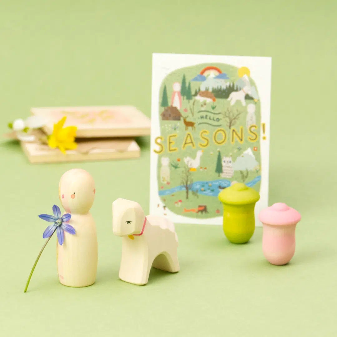 Vor einem hellgrünen Hintergrund ist das limitierte „Hello Spring“ Jahreszeitenset Frühling von Ostheimer und Grapat zu sehen: eine helle Holzfigur, ein weißes Lamm, zwei pastellfarbene Holzformen in Rosa und Grün sowie eine violette Blüte. Im Hintergrund steht eine illustrierte Jahreszeiten-Karte.