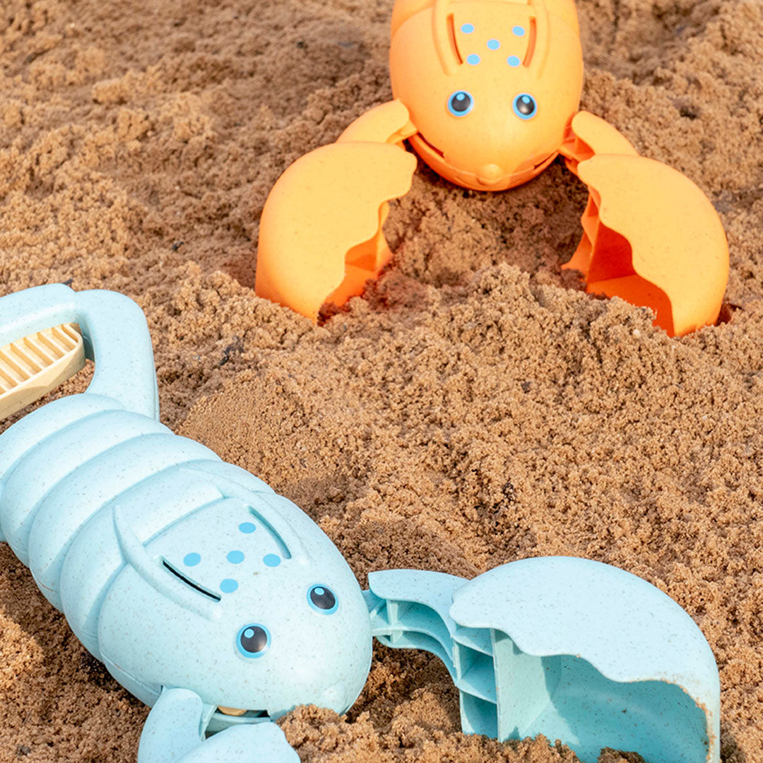Ein hellblauer und ein orangener Hummer aus einem Kunststoff-Weizenstroh-Gemisch auf Sand platziert, haben einen Griff der zudrückbar ist. Die Scheren lassen somit öffnen und schließen. Sie haben einen oval geformten Körper, der mit zwei Augen und fünf blauen Punkten versehen ist. Beide sind jeweils im Anschnitt zu sehen.