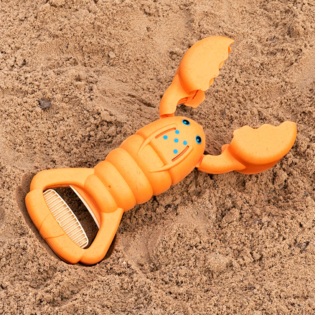 Ein orangener Hummer aus einem Kunststoff-Weizenstroh-Gemisch auf Sand platziert, hat einen Griff der zudrückbar ist. Die Scheren lassen somit öffnen und schließen. Er hat einen oval geformten Körper, der mit zwei Augen und fünf blauen Punkten versehen ist.