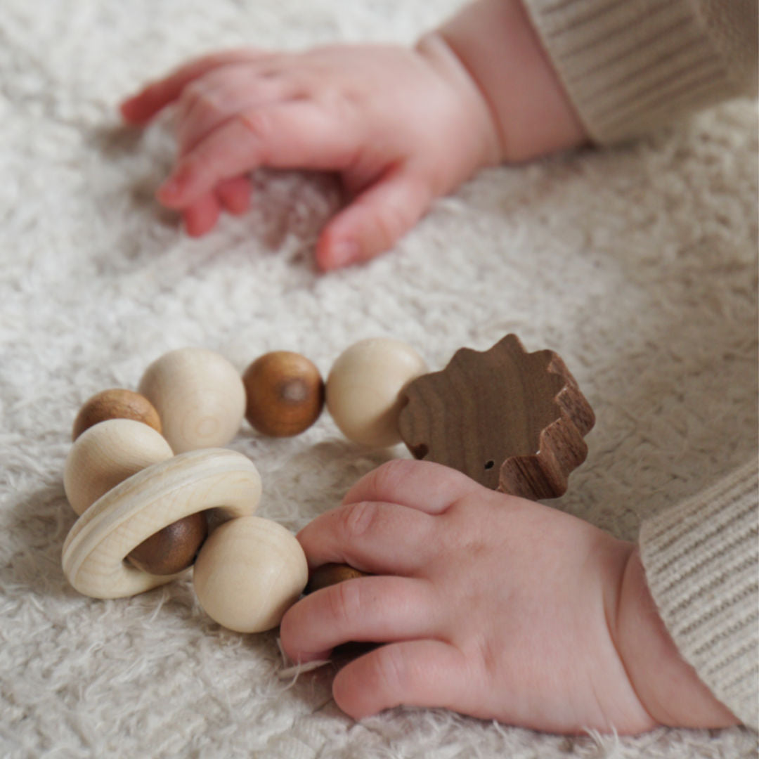 Ein Greifling aus zweifarbigem Holz mit bewegbarem Ring, Perlen und einem Igel von Holztiger, der auf einem Teppich liegt und von einem Kleinkind in der linken Hand liegt.