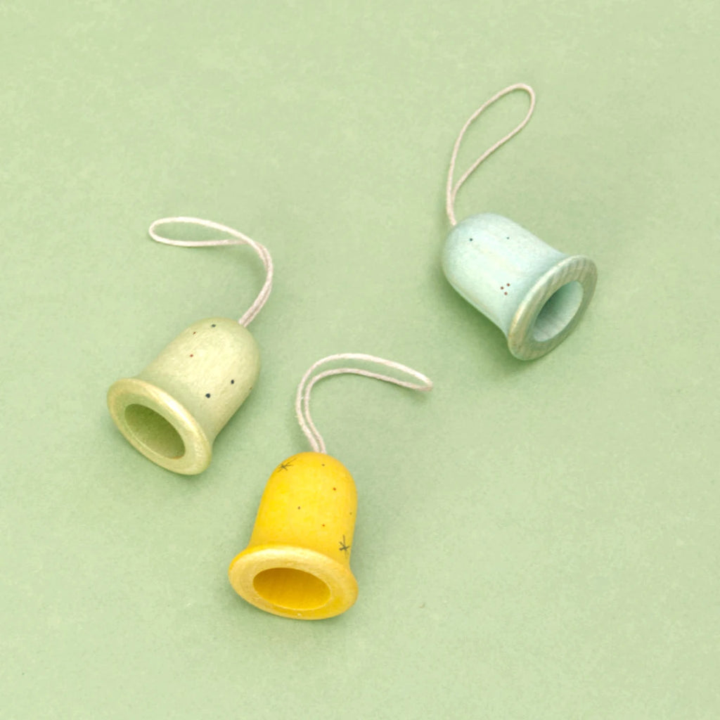 Die drei Glocken der Serie Jingle Joy Bells von den Grapat Ornaments liegen auf einem grünen Untergrund. Die Weihnachtsbaumanhänger sind aus Holz gefertigt und in Gelb, zartem Blau und feinem Grün gestaltet. Sie sind mit Sternen, Sprenkeln und weiteren farblichen Akzenten sowie goldenen Verzierungen geschmückt.