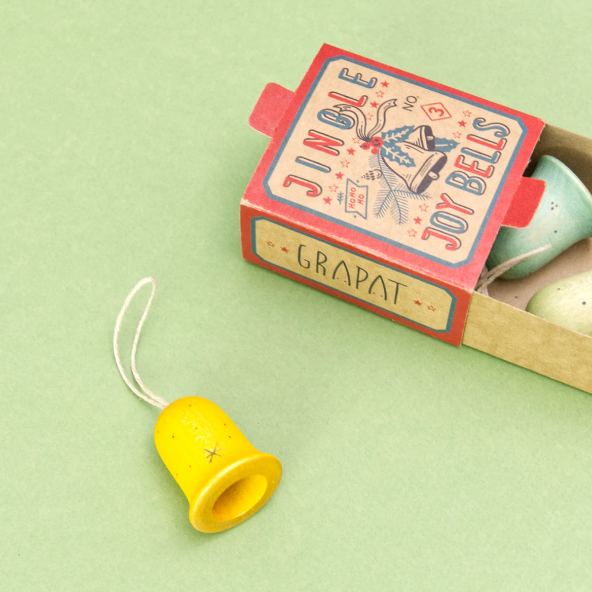 Die Verpackung der Serie Jingel Joy Bells von den Grapat Ornaments liegt offen auf einem grünen Untergrund und beinhaltet zwei von den drei bunten Glocken Weihnachtsbaumanhängern. Der dritte Anhänger liegt außerhalb der Schachtel und ist gelb mit dezentem Pünktchenmuster.