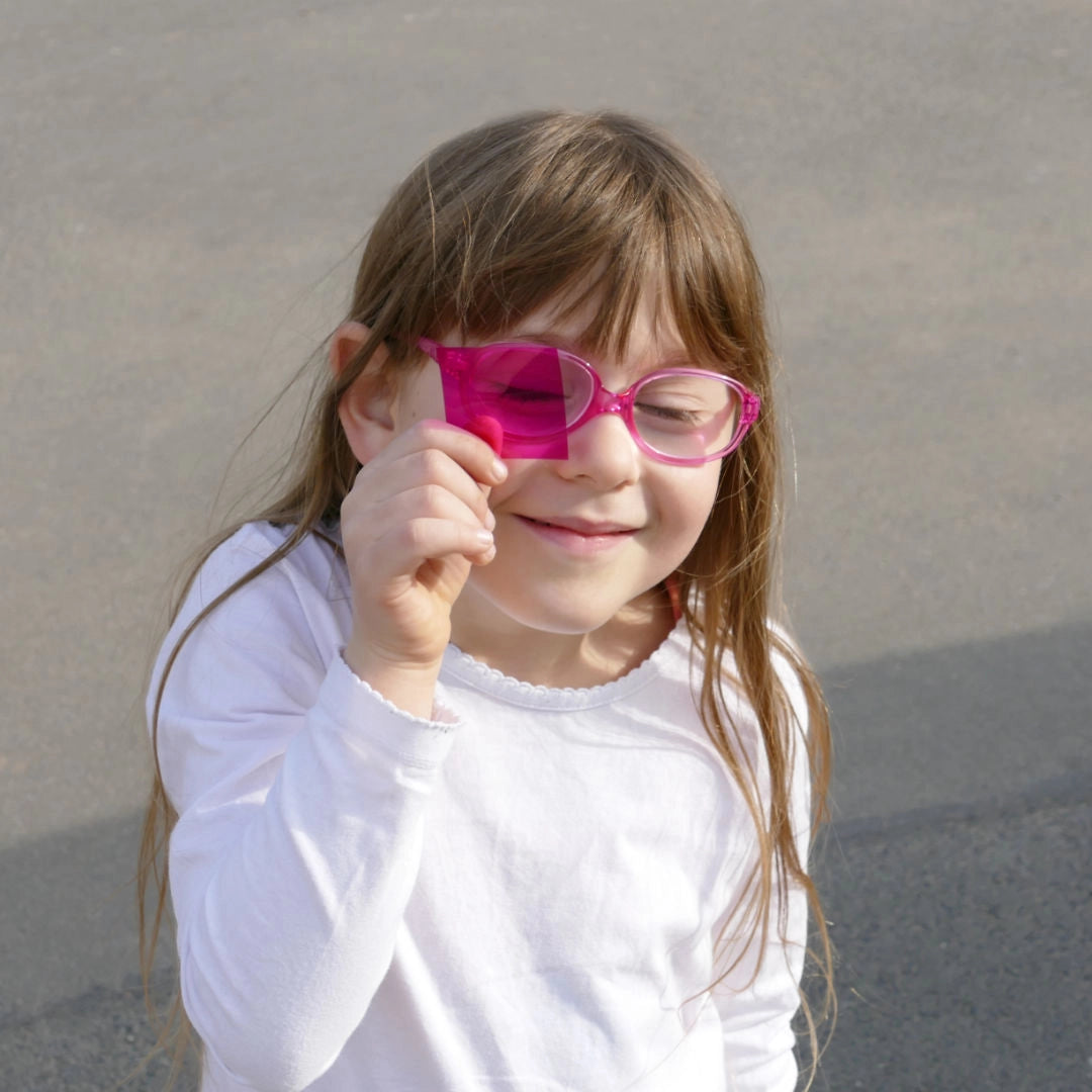Ein Mädchen hält eine pinke Farbkarte vor ihr Gesicht. Sie trägt eine pinke Brille, hat die Augen zusammengekniffen und lächelt freundlich. Das humorvolle Foto steht sinnbildlich für das Entdecken und greift dabei spielerisch das Thema: "Die Augen auf etwas richten" auf.