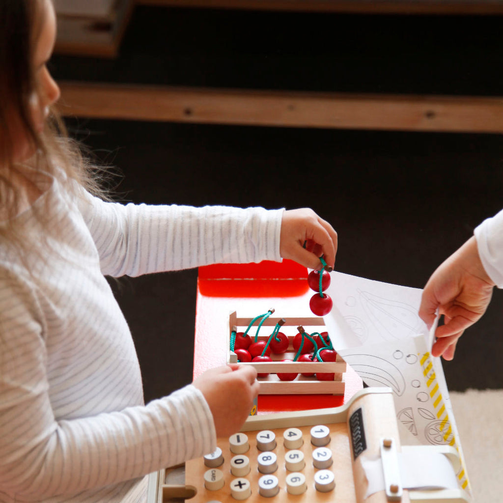 Spielsituation im Kaufladen, Spielkirschen werden in Papier Obsttüte gefüllt. Auf der Theke stehen Obststeige mit Holzkirschen sowie Spielkasse aus Holz.