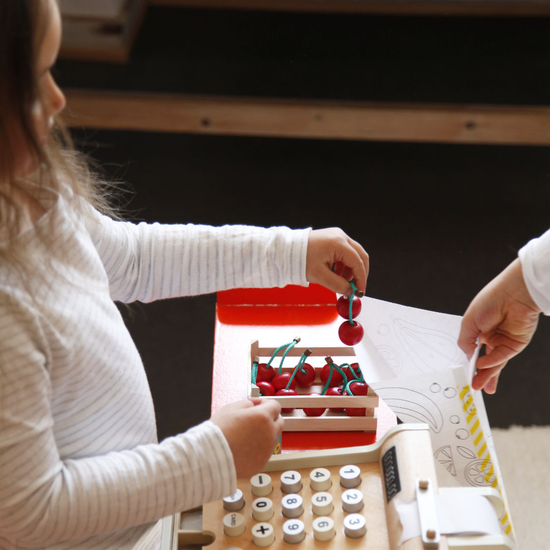 Spielsituation im Kaufladen, Spielkirschen werden in Papier Obsttüte gefüllt. Auf der Theke stehen Obststeige mit Holzkirschen sowie Spielkasse aus Holz.
