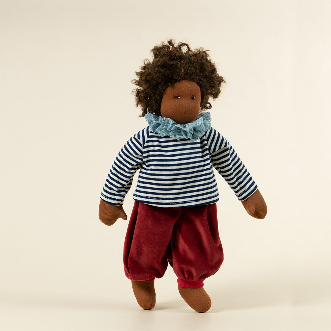Nanchen Puppe mit dunkler Haut und dunkeln Haaren stehend. Sie trägt ein dunkelblau-weiß gestreiftes Oberteil mit Kragen und eine rote Hose.