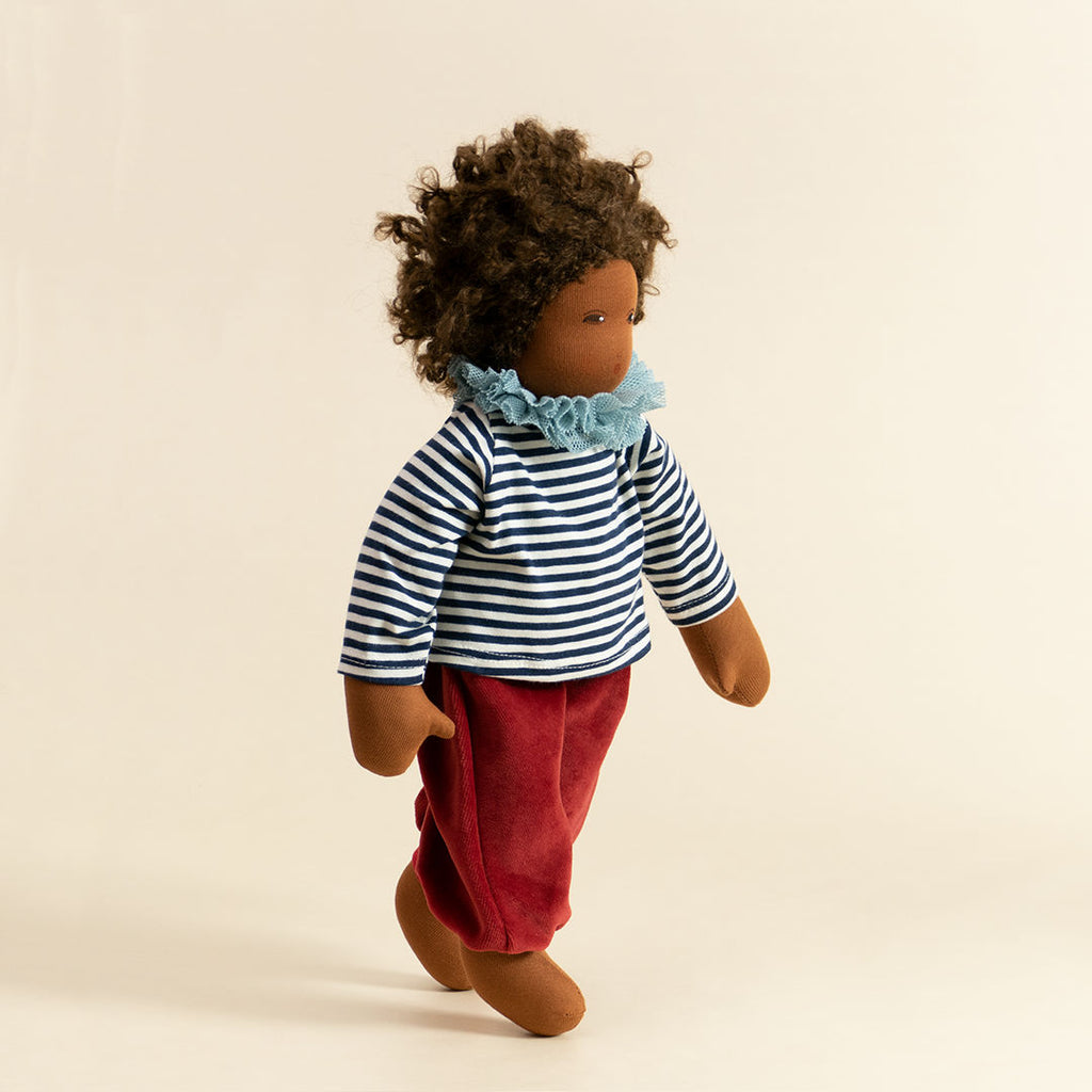 Nanchen Puppe mit dunkler Haut und dunkeln Haaren stehend. Sie trägt ein dunkelblau-weiß gestreiftes Oberteil mit Kragen und eine rote Hose.