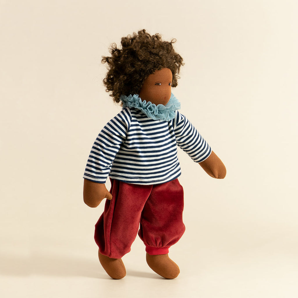 Nanchen Puppe mit dunkler Haut und dunkeln Haaren stehend. Sie trägt ein dunkelblau-weiß gestreiftes Oberteil mit Kragen und eine rote Hose.