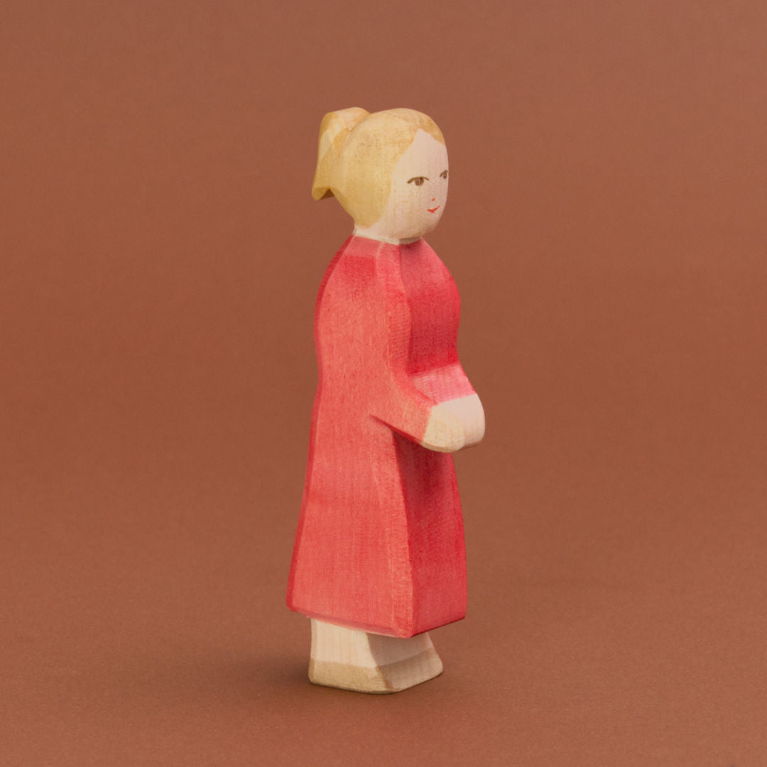 Eine Mutter aus Holz von der Marke Ostheimer mit heller Hautfarbe, blonden Haaren, die zu einem Pferdeschwanz gebunden sind, und hellbraunen Augen ist leicht eingedreht stehend und nach rechts vorne schauend, zu sehen. Ihr Mund und die Nase sind rot bemalt und ihre beiden Arme hält sie nach vorne zusammen. Sie trägt ein pinkes, langes Kleid und hellbraune Schuhe.