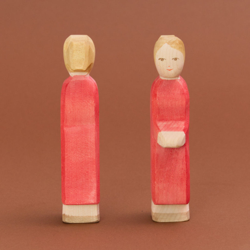 Zwei blonde Mütter aus Holz mit heller Hautfarbe und pinken Kleidern sind parallel nebeneinanderstehend zu sehen. Die rechte Mutter schaut nach vorne und ist mit dem Gesicht zu sehen, die linke Mutter schaut nach hinten und ist nur von hinten zu sehen.