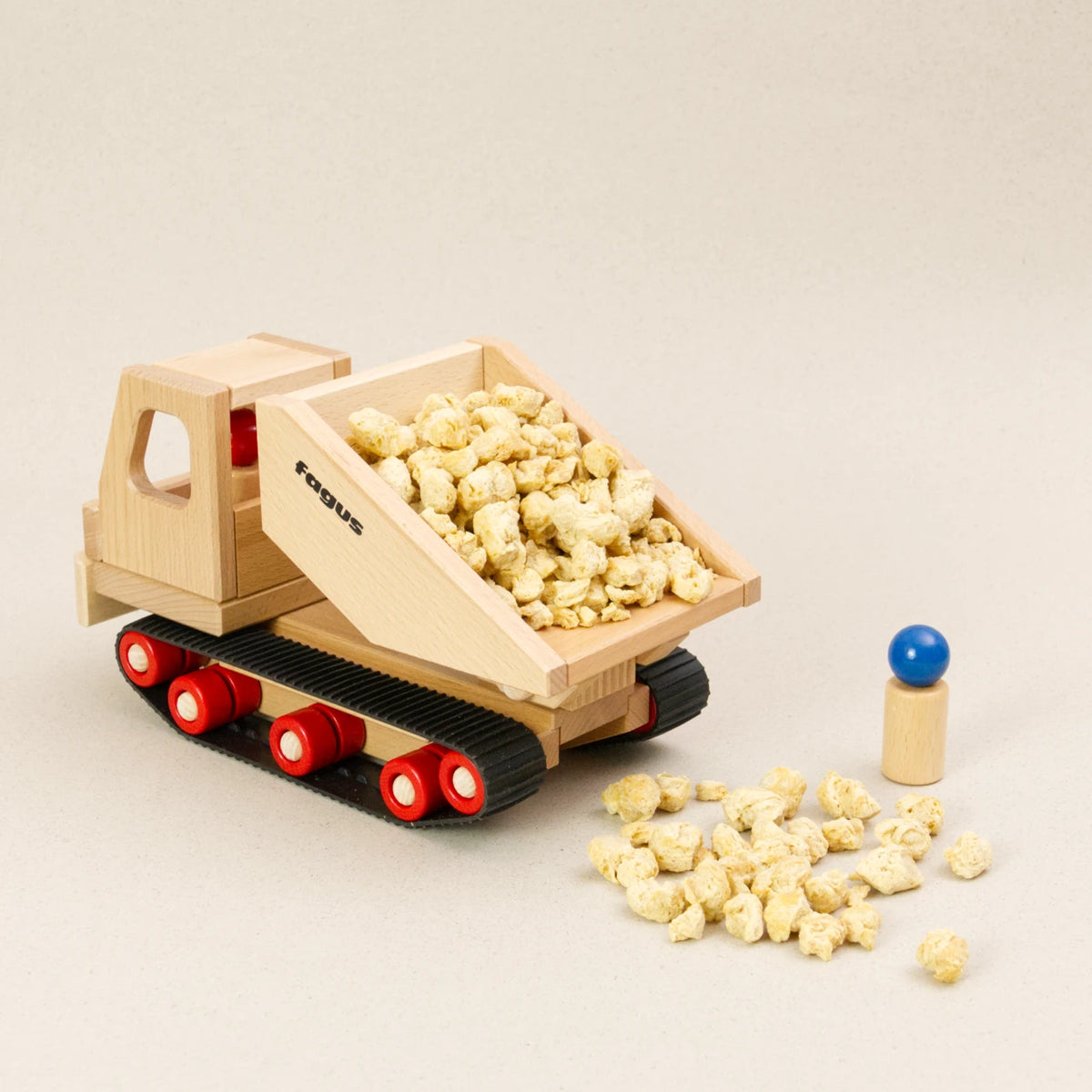Der fagus Raupen Dumper ist aus naturbelassenem Buchenholz, hat eine kantige Lademulde und fährt auf Gummiketten. Ansicht von hinten, die Ladefläche ist leicht gekippt, eine Holzfigur steht daneben und überwacht das Entladen des Schuttguts.