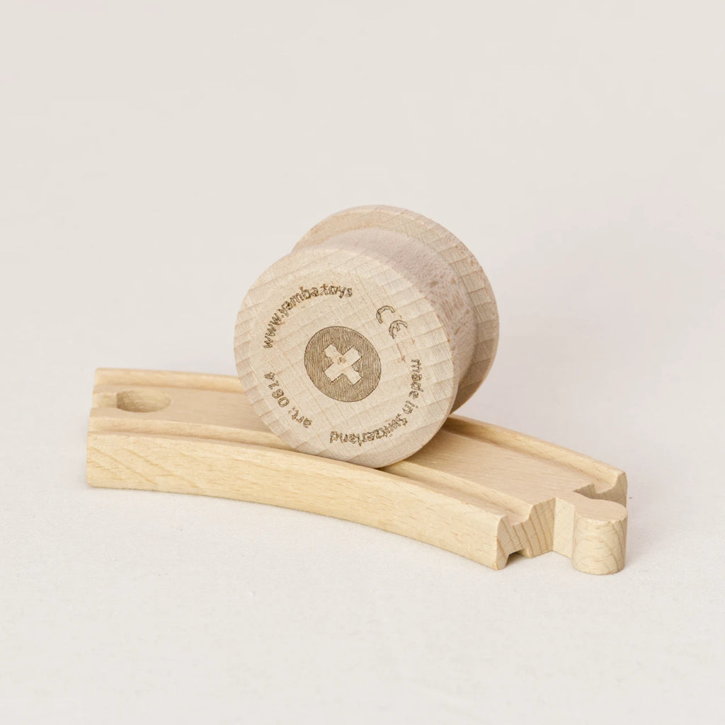 Zu sehen ist die Holzwalze Rolli von Yamba auf einer Spielzeug-Holzschiene. Der Rolli hat die Form eines Zylinders mit leicht überstehenden Enden, um in der Schiene laufen zu können (BRIO®, IKEA®, Eichhorn®, Hape® usw.). Er besteht aus naturbelassener Schweizer Buche und ist aus einem Stück gefertigt, ohne Leim und Lack. Auf der flachen Seite ist mittig ein Kreuz in einem Kreis zu sehen. Am Rand stehen Informationen wie Herstellungsort und Artikelnummer.