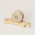 Zu sehen ist die Holzwalze Rolli von Yamba auf einer Spielzeug-Holzschiene. Der Rolli hat die Form eines Zylinders mit leicht überstehenden Enden, um in der Schiene laufen zu können (BRIO®, IKEA®, Eichhorn®, Hape® usw.). Er besteht aus naturbelassener Schweizer Buche und ist aus einem Stück gefertigt, ohne Leim und Lack. Auf der flachen Seite ist mittig ein Kreuz in einem Kreis zu sehen. Am Rand stehen Informationen wie Herstellungsort und Artikelnummer.
