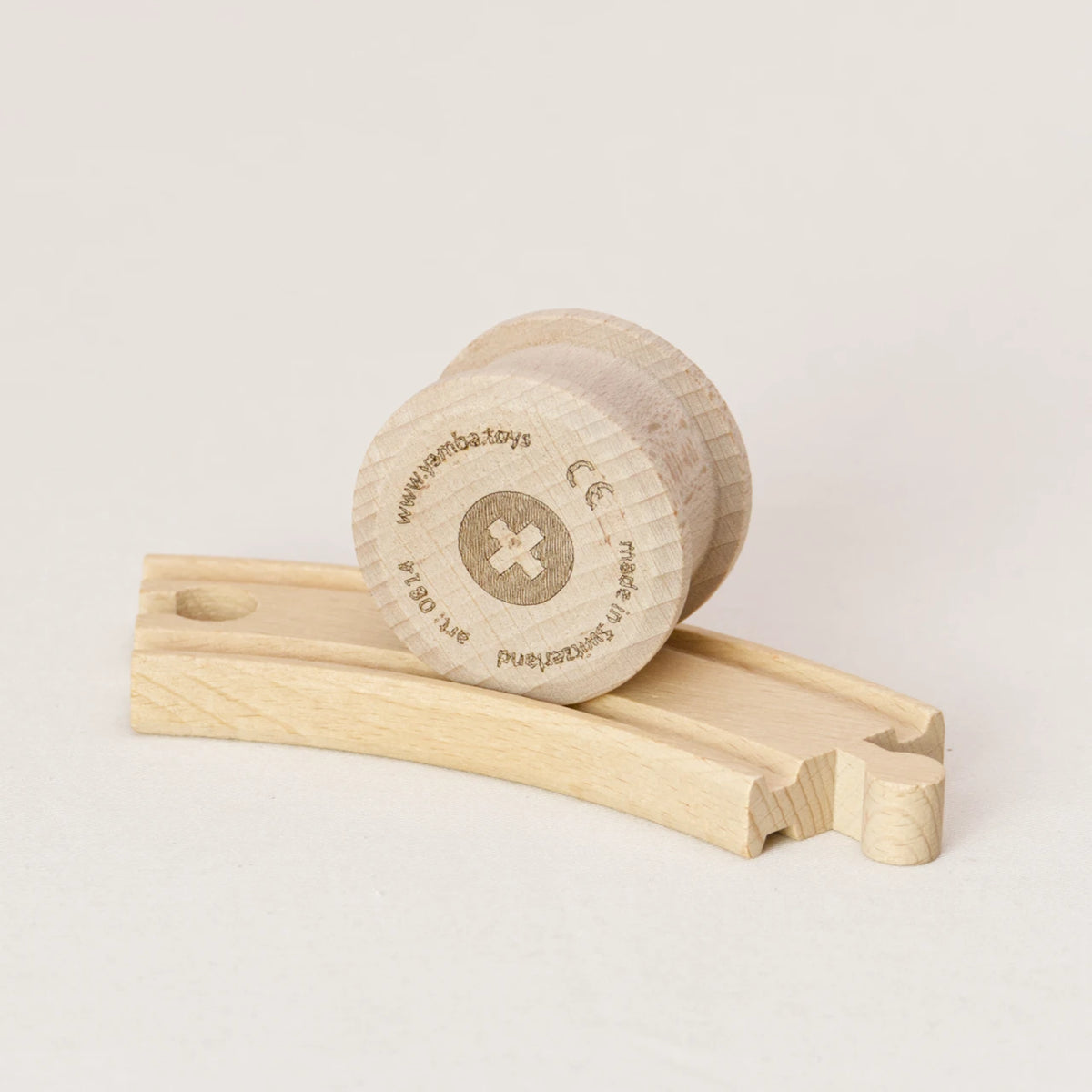 Zu sehen ist die Holzwalze Rolli von Yamba auf einer Spielzeug-Holzschiene. Der Rolli hat die Form eines Zylinders mit leicht überstehenden Enden, um in der Schiene laufen zu können (BRIO®, IKEA®, Eichhorn®, Hape® usw.). Er besteht aus naturbelassener Schweizer Buche und ist aus einem Stück gefertigt, ohne Leim und Lack. Auf der flachen Seite ist mittig ein Kreuz in einem Kreis zu sehen. Am Rand stehen Informationen wie Herstellungsort und Artikelnummer.