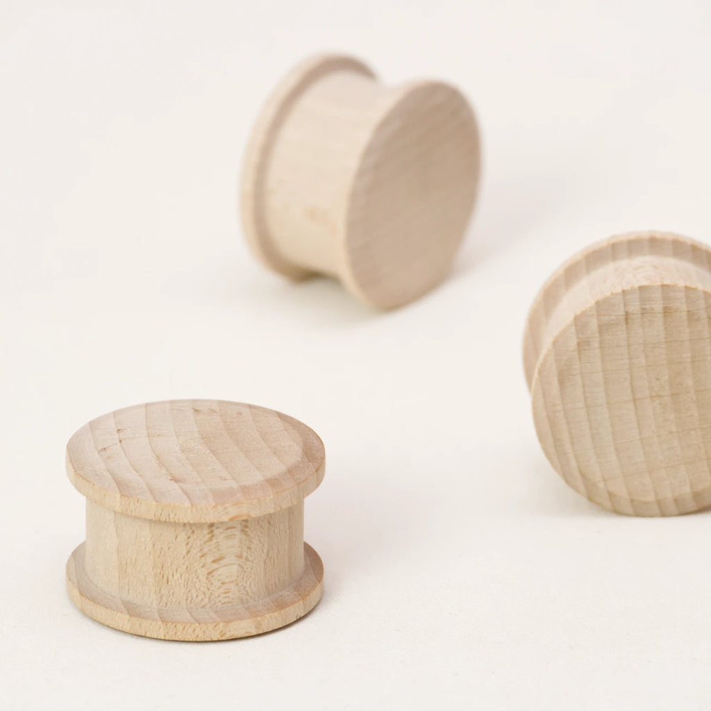 Zu sehen sind drei Rolli-Holzwalzen von Yamba in unterschiedlichen Positionen. Die Rollis haben die Form eines Zylinders. Einer steht auf der flachen Unterseite, die beiden anderen auf der gebogenen Mantelfläche. Einer ist in der Seitenansicht zu sehen, der andere steht schräg. Die Rollis bestehen aus naturbelassener Schweizer Buche und sind aus einem Stück gefertigt. Auf Leim und Lack wird verzichtet.