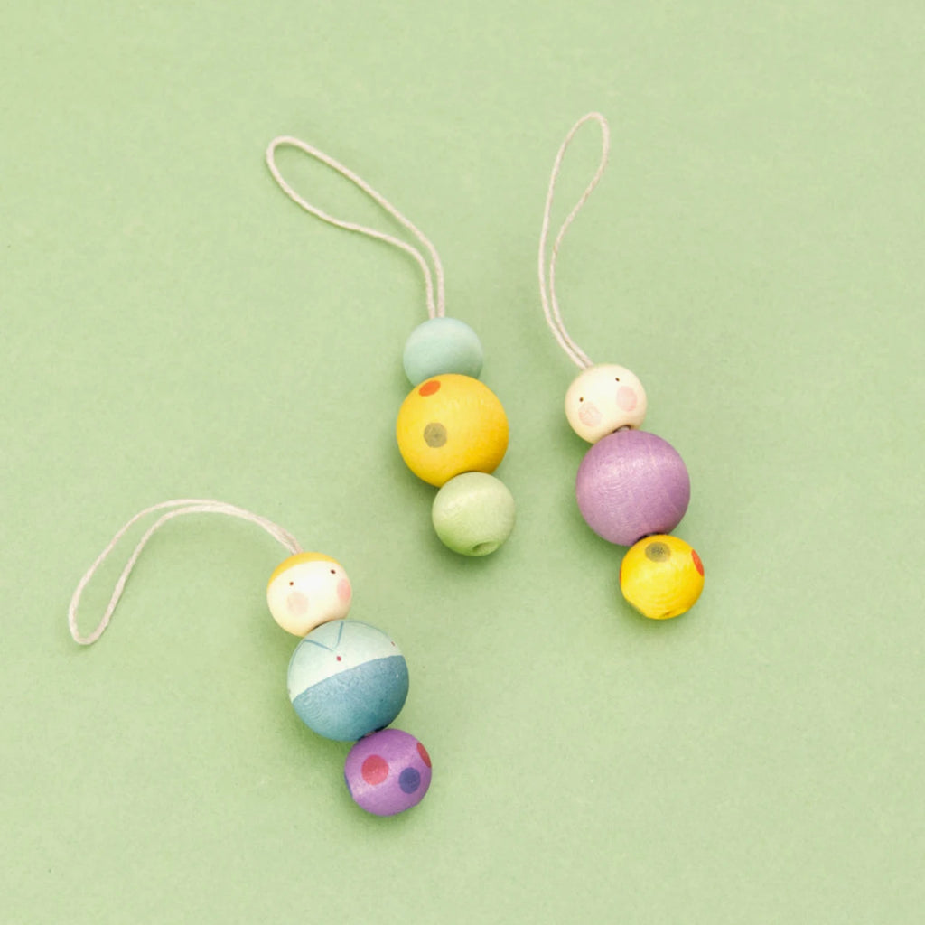 Die drei Figuren der Serie Snowy Buddy von den Grapat Ornaments liegen auf einem grünen Untergrund. Alle Weihnachtsbaumanhänger bestehen aus drei Holzkugeln. Der mittig platzierte Anhänger ist bunt gemustert, während die beiden anderen Figuren ein Gesicht haben. Die Anhänger sind in Violett, Gelb, Blau und Grün gestaltet, mit weiteren farblichen Akzenten.