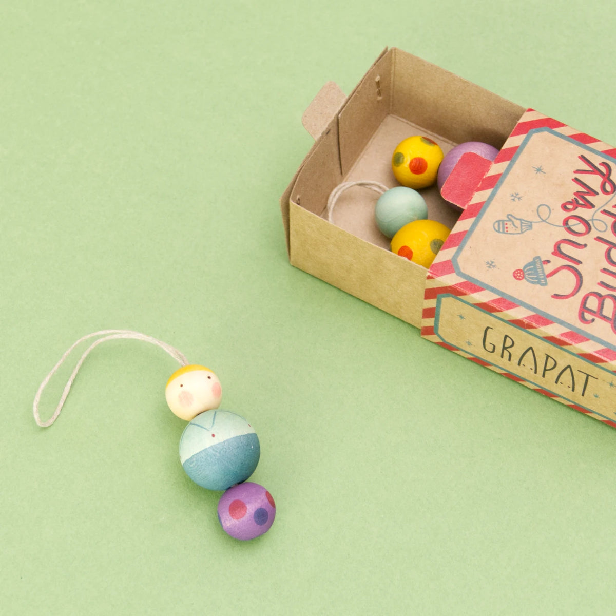 Die Verpackung der Serie Snowy Buddy von den Grapat Ornaments liegt offen auf einem grünen Untergrund und beinhaltet zwei von den drei bunt gemusterten Weihnachtsbaumanhängern. Der dritte Anhänger liegt außerhalb der Schachtel und hat ein Gesicht und grün, blau, violette Kugeln mit Musterung.