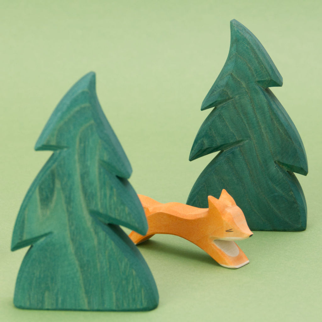 Ein Fuchs aus Holz von der Marke Ostheimer läuft von links nach rechts durch zwei dunkelgrün bemalten Tannen aus Holz, ebenfalls von der Marke Ostheimer.