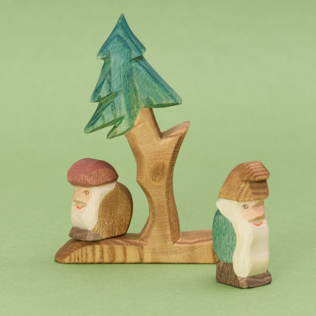 Eine Figurengruppe der Märchenwelt von Ostheimer mit zwei Tannenzwergen und einem Bäumchen. Die hölzernen Spielfiguren zeigen einen kleineren Zwerg, der einen Buckel macht, ein braunes Mäntelchen und eine rote Mütze trägt und einen kurzen Vollbart hat. Er steht auf einem Baumstumpf, schaut nach links und lächelt. Der größere Zwerg steht aufrecht, trägt ein grünes Gewand und eine braune Zipfelmütze und hat einen langen weißen Bart. Die Tanne hat einen längeren Stamm und einen vorstehenden Ast.