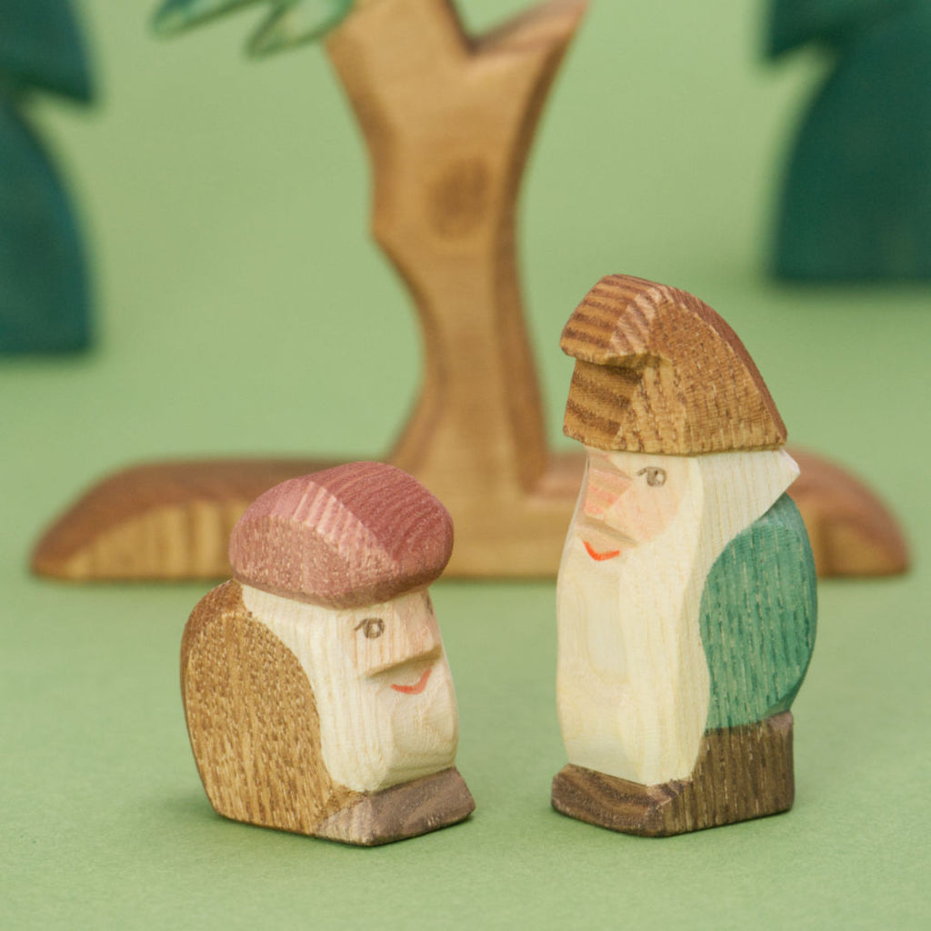 Zwei hölzerne Zwergenfiguren der Marke Ostheimer vor einer Tanne. Die Holzmaserung der Figuren ist in der Detailaufnahme gut erkennbar. Der kleinere Zwerg macht einen Buckel, trägt ein braunes Mäntelchen, eine rote Mütze und hat einen kurzen Vollbart. Der größere Zwerg steht aufrecht, trägt ein grünes Gewand und eine braune Zipfelmütze, mit einem langen weißen Bart. Im Hintergrund sind unscharf weitere Bäume zu sehen.