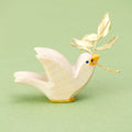 Eine hölzerne Taube der Marke Ostheimer, die nach rechts schaut. Die flach ausgesägte Figur zeigt die Silhouette eines fliegenden Vogels, die Kanten sind leicht abgerundet. Die Taube ist weiß lasiert, hat einen gelben Schnabel und gelbe Füße, die Augen bestehen aus kleinen blauen Punkten. Im Schnabel steckt ein sehr kleines Ästchen mit winzigen goldenen Blättern.
