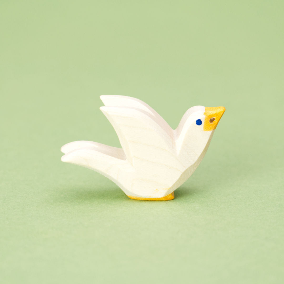Eine hölzerne Taube der Marke Ostheimer, die nach rechts schaut. Die flach ausgesägte Figur zeigt die Silhouette eines fliegenden Vogels, die Kanten sind leicht abgerundet. Die Taube ist weiß lasiert, hat einen gelben Schnabel und gelbe Füße, die Augen bestehen aus kleinen blauen Punkten. Im Schnabel befindet sich ein Loch, durch das ein kleines Ästchen gesteckt werden könnte.